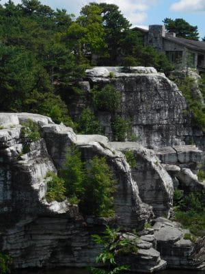 A stone building sits atop tall, white rocky cliffs surrounded by dense green trees, under a partly cloudy blue sky.