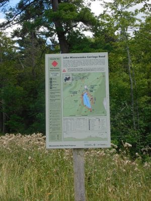 A trail sign for Lake Minnewaska Carriage Road stands in a grassy area with trees in the background. The sign features a map, trail information, and safety guidelines for hikers at Minnewaska State Park Preserve.