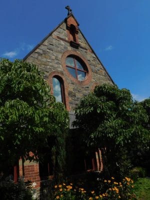 A stone church with arched windows and a cross on top, partly shaded by green trees and surrounded by colorful flowers, stands near The Abbey Inn under a bright blue sky.