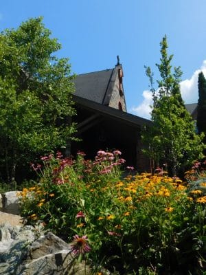 A garden with blooming purple and yellow flowers, lush green trees, and rocks in the foreground. In the background, The Abbey Inn church stands with a pointed tower and steep, dark roofs under a bright blue sky with clouds.