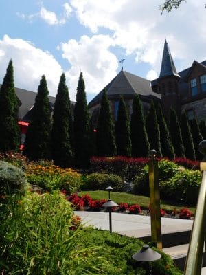 A beautifully landscaped garden with colorful flowers and lush greenery leads up to The Abbey Inn, a large stone building with pointed rooftops and tall, narrow evergreen trees under a partly cloudy sky.