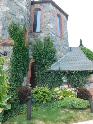 The Abbey Inn is a stone building with tall, narrow windows and reddish brick accents, partially covered by green ivy. Lush shrubs grow near the base, with a shingled roof visible on the right side.