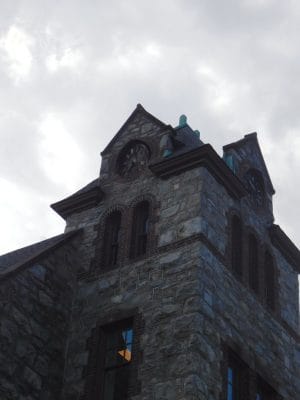 A stone clock tower with arched windows stands against a cloudy sky at The Abbey Inn. The clock faces are visible on two sides, and part of the building’s roof is also shown.