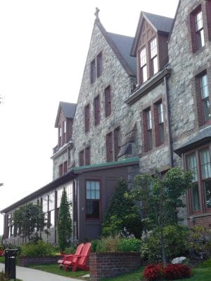 The Abbey Inn is a large stone building with steep gabled roofs and many windows, surrounded by landscaped gardens, trees, and red Adirondack chairs on a lawn, all under a bright sky.