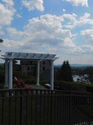 A white pergola with red chairs at The Abbey Inn overlooks a scenic river, distant shoreline, and cloudy sky. Lush greenery and a stone building sit beside a black iron railing in the foreground.