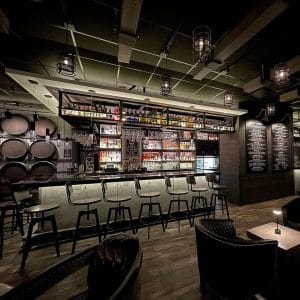 A dimly lit bar with high chairs, shelves stocked with bottles, hanging glasses, and barrel decor on the left. Shaker & Vine menu boards are displayed on the wall, creating a cozy, modern atmosphere.