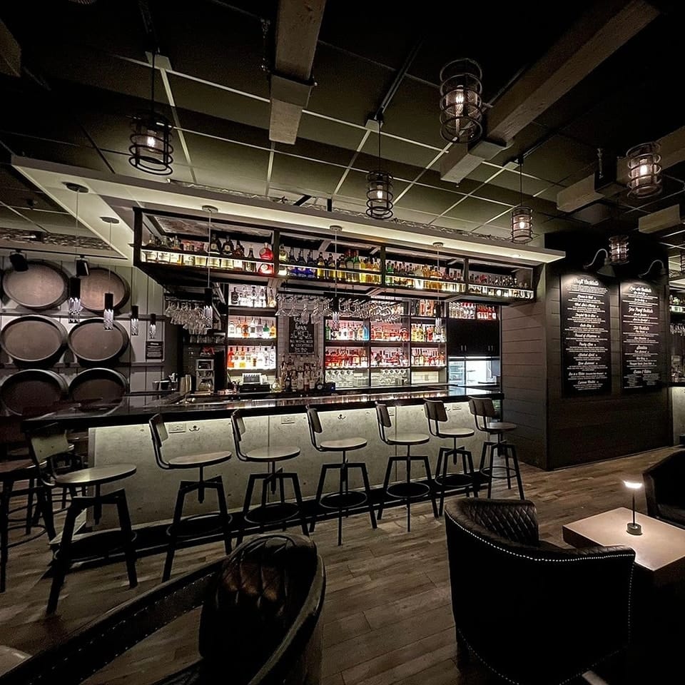 A dimly lit bar with high chairs, shelves stocked with bottles, hanging glasses, and barrel decor on the left. Shaker & Vine menu boards are displayed on the wall, creating a cozy, modern atmosphere.