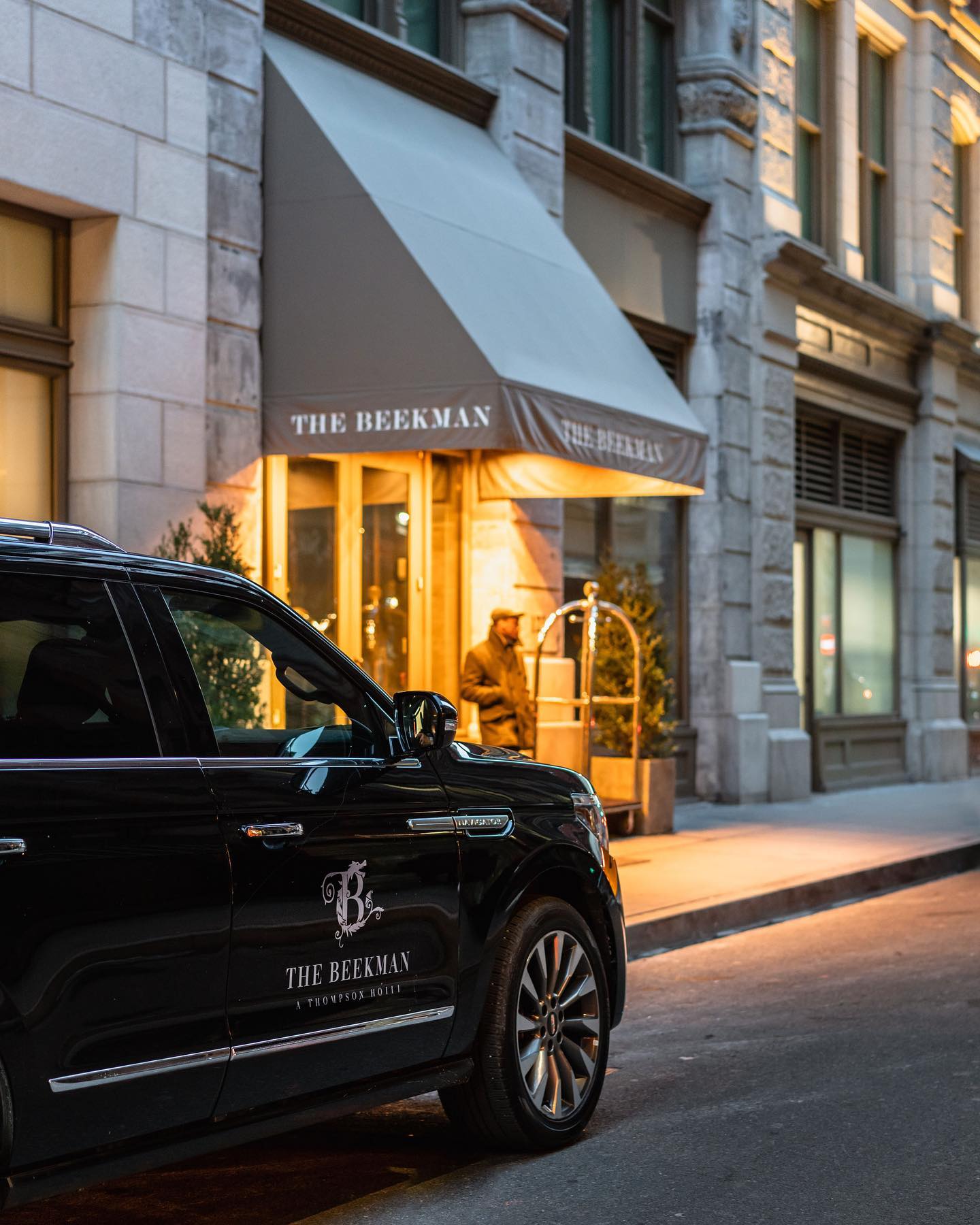 A black SUV with The Beekman Hotel logo is parked in front of the entrance as a doorman stands by a luggage cart under the illuminated awning on a city street at dusk.