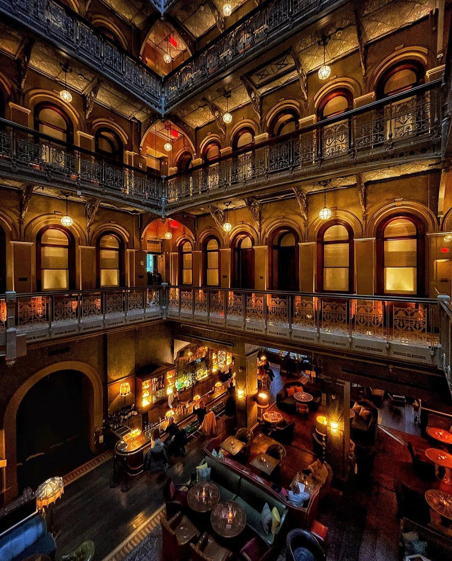 A warmly lit, multi-story interior of The Beekman Hotel features ornate railings, arched windows, cozy seating areas, and a central bar on the ground floor, creating an inviting, vintage atmosphere.
