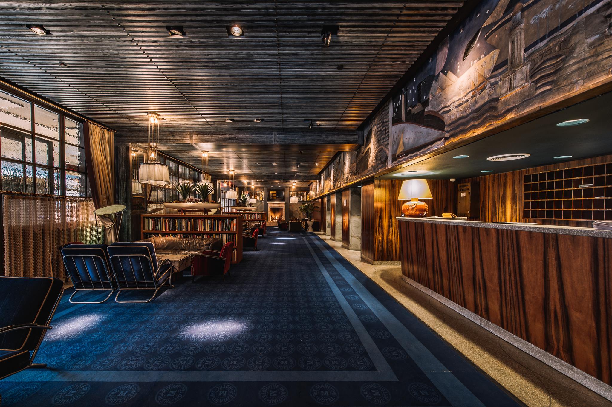 The spacious Maritime Hotel lobby features a wooden ceiling and walls, a long patterned carpet, vintage furniture, large lamps, lush plants, and a welcoming reception desk with warm lighting and decorative art on the walls.