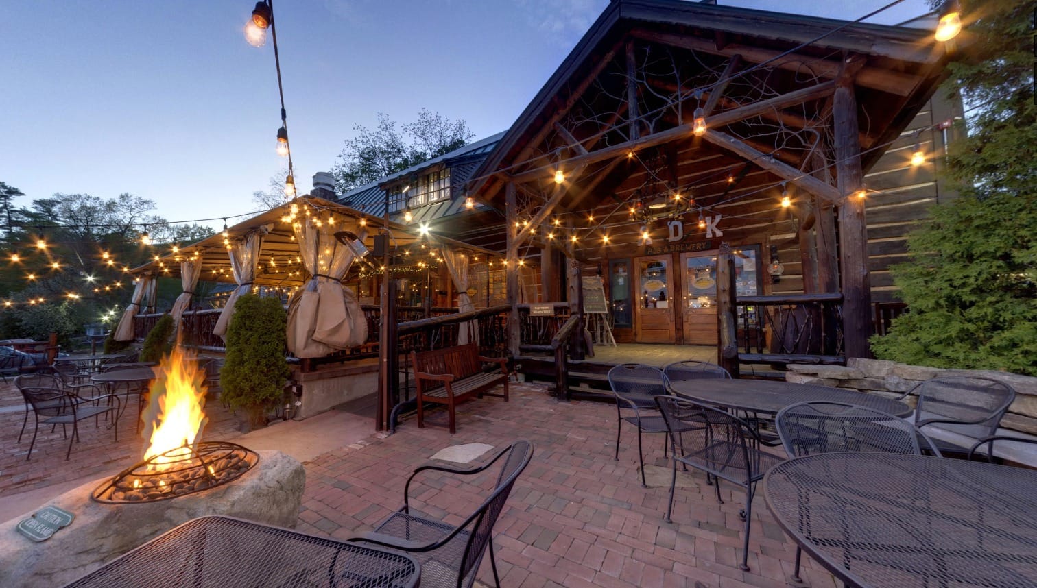 Outdoor dining area at Adirondack Pub & Brewery with empty tables and chairs on a brick patio, string lights overhead, a fire pit burning, and a rustic wooden building in the background at dusk.