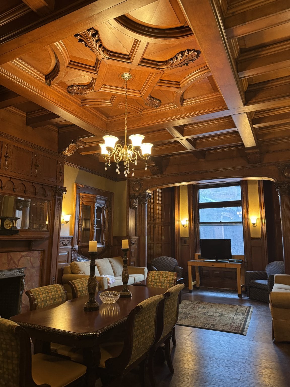 A warmly lit room with ornate wood paneling and an intricate coffered ceiling features a dining table with chairs, a chandelier, sofas, armchairs, a fireplace, and a TV near a large window.