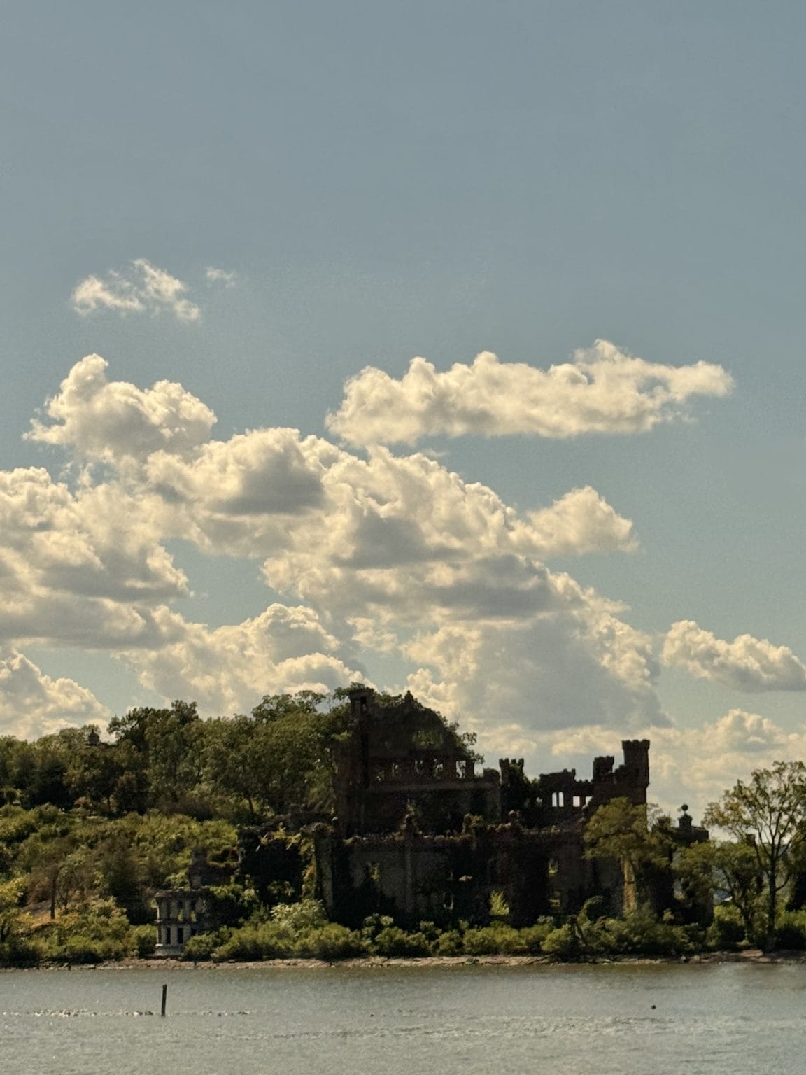 Ruins of an old castle-like building covered in greenery sit by the water’s edge, surrounded by trees, under a sky filled with fluffy clouds and soft sunlight.