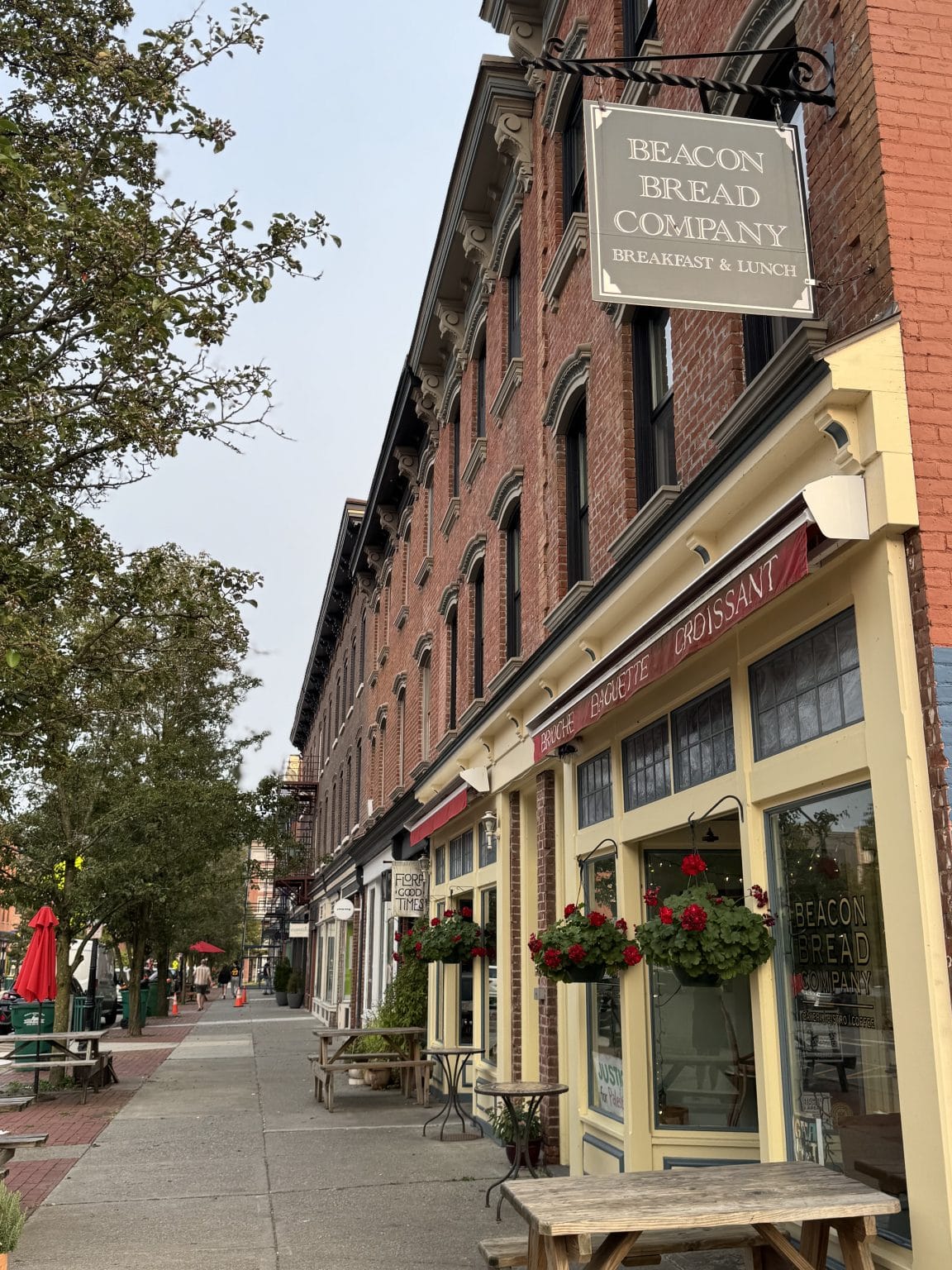 A street view of historic brick buildings with storefronts and outdoor seating. “Beacon Bread Company” sign hangs above a bakery with large windows, red awnings, and flower boxes. Trees and lampposts line the sidewalk.