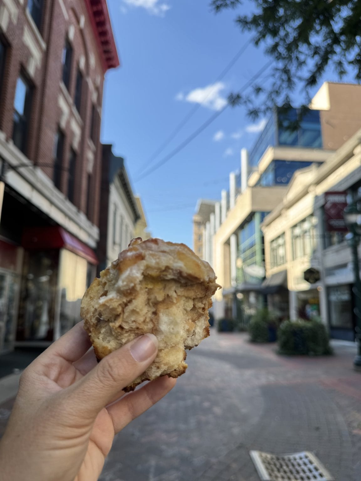 A hand holds a partially eaten pastry on a brick-paved city street lined with shops and buildings under a blue sky with scattered clouds.