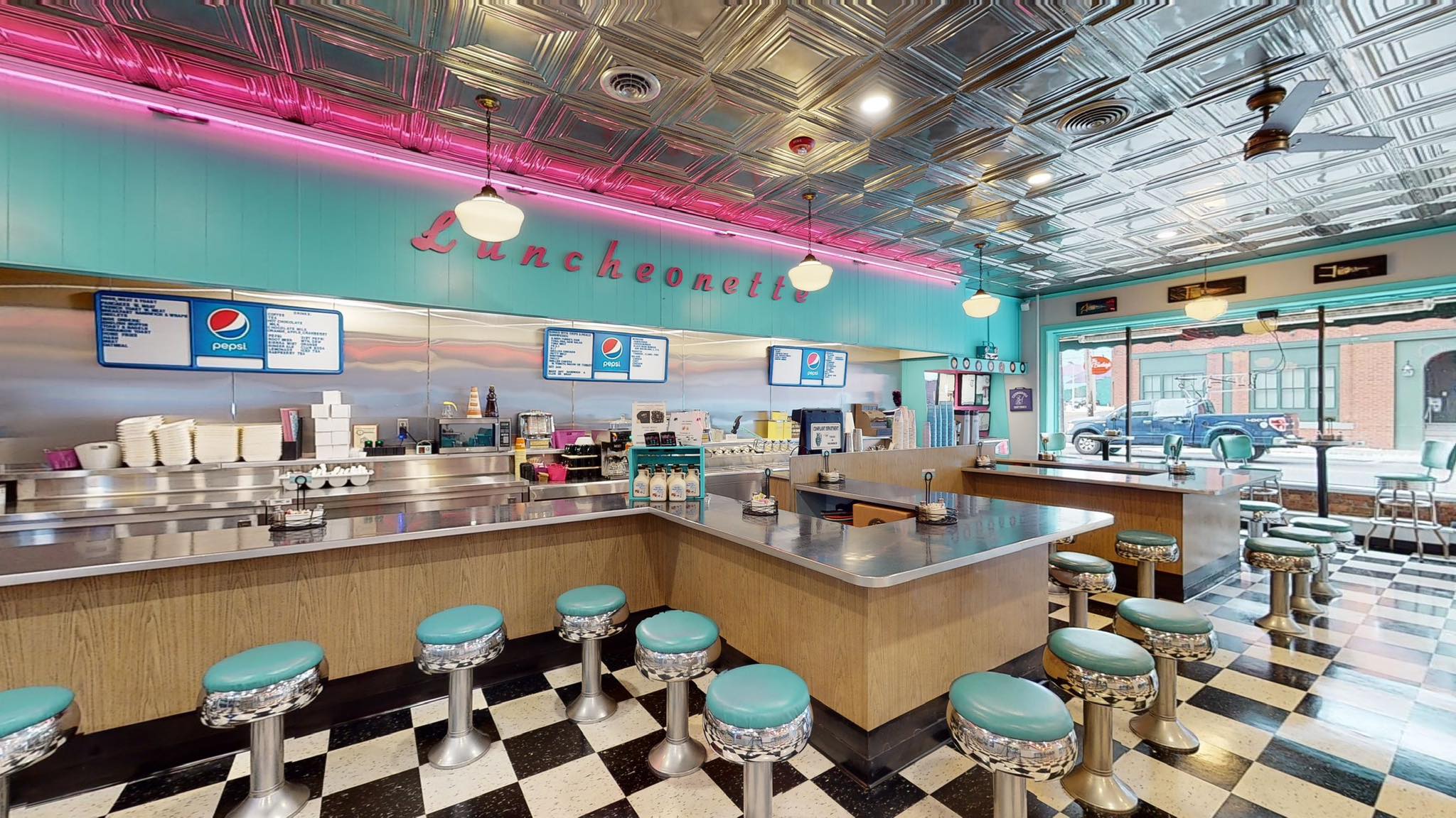 A retro diner, Burleigh’s Luncheonette, with teal and pink decor, chrome accents, checkered black-and-white floor, teal-cushioned stools, a long counter, and large windows. Menus and soda signs hang on the back wall.