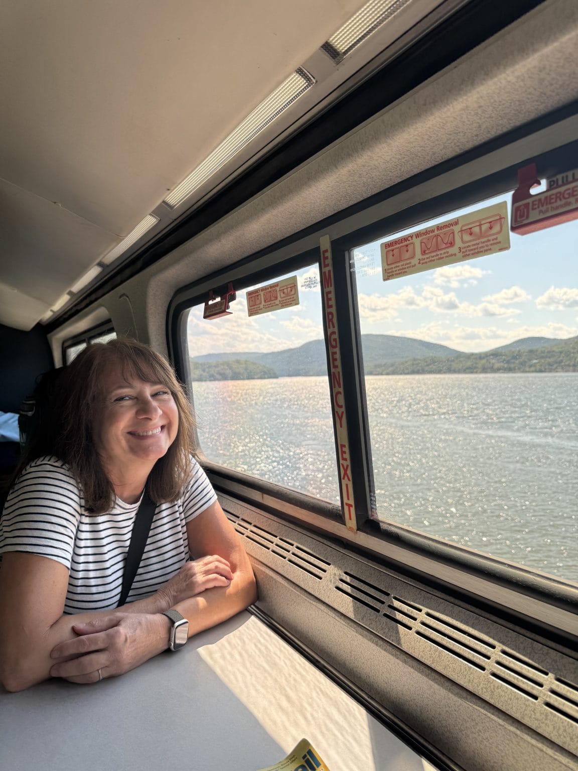 A woman with shoulder-length brown hair and a striped shirt smiles while sitting at a table by a train window, with a scenic view of a lake and distant hills under a blue sky outside.