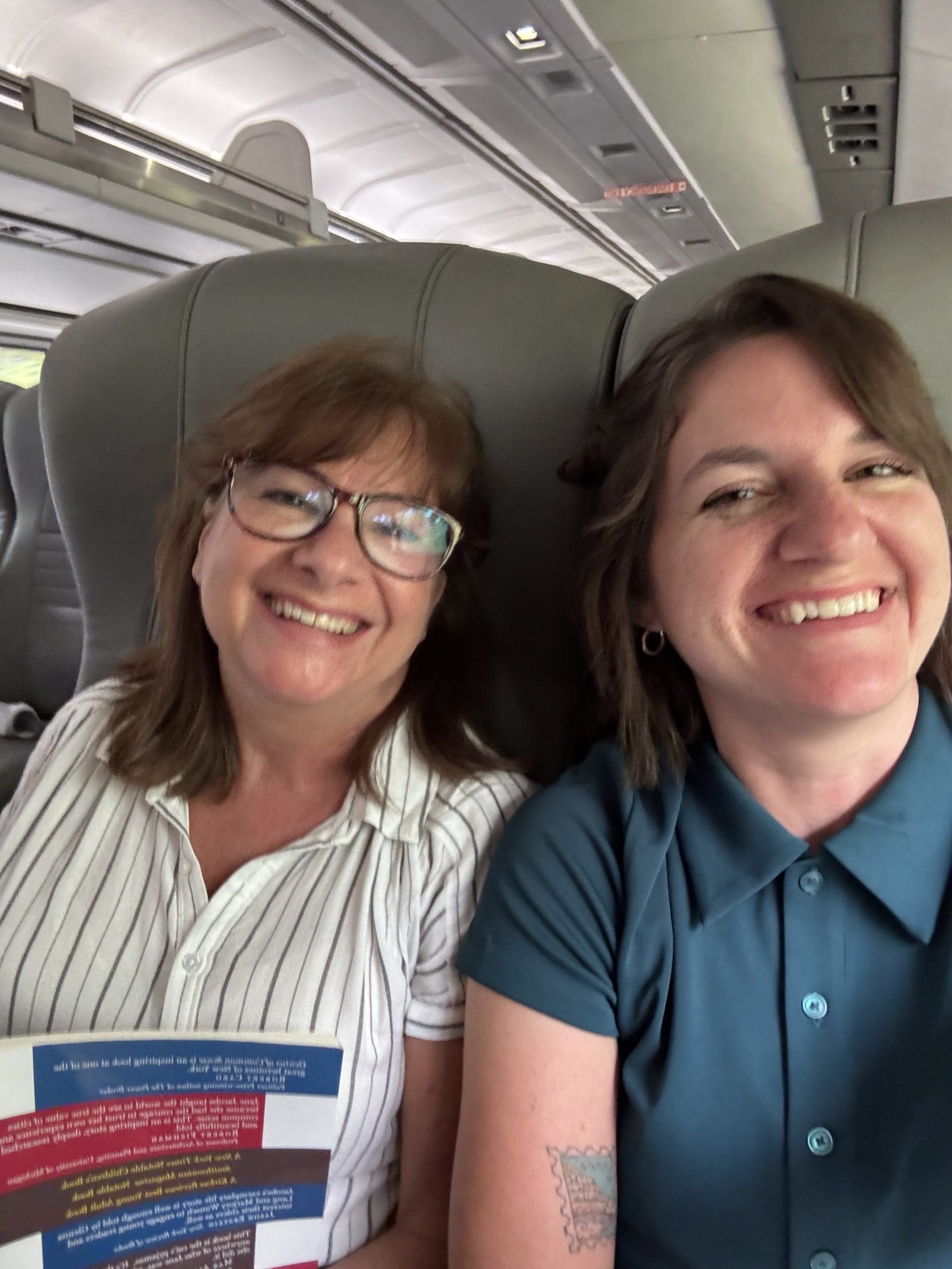 Two women smiling and sitting next to each other on a train, one holding a colorful booklet, both in collared shirts. They're ready to explore Upstate New York, surrounded by grey train seats and the sleek train interior ceiling.