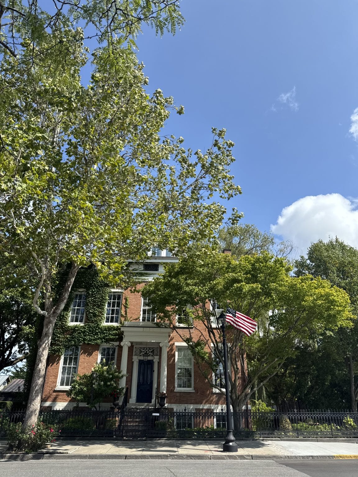 A red brick house partly covered in ivy sits behind a black metal fence. An American flag is displayed on a lamppost in front, and trees with green leaves partially obscure the house under a clear, sunny sky.