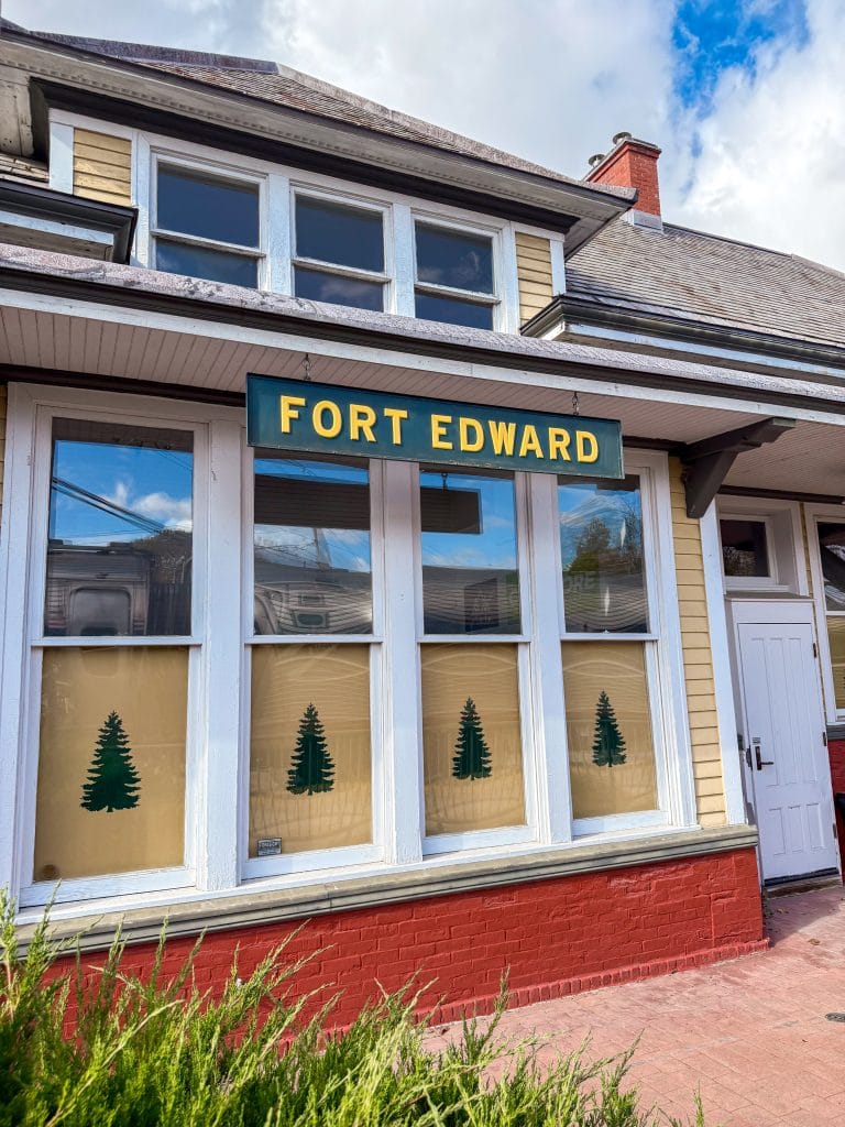 A yellow building with a green and yellow sign reading Fort Edward above windows decorated with pine tree silhouettes. Shrubs are in the foreground under a partly cloudy sky near the Miss New York Lake George pageant route.