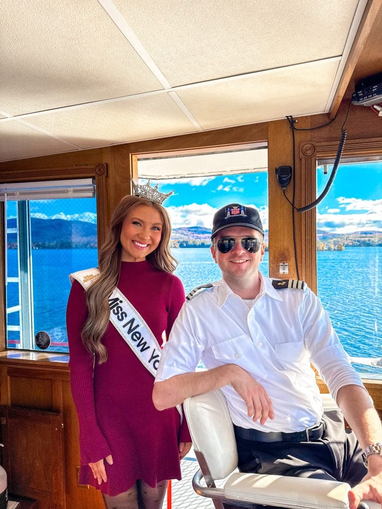 A woman wearing a crown and a Miss New York Lake George sash stands smiling next to a man in a captain's uniform and sunglasses inside a boat, with blue water and sky visible through the windows.