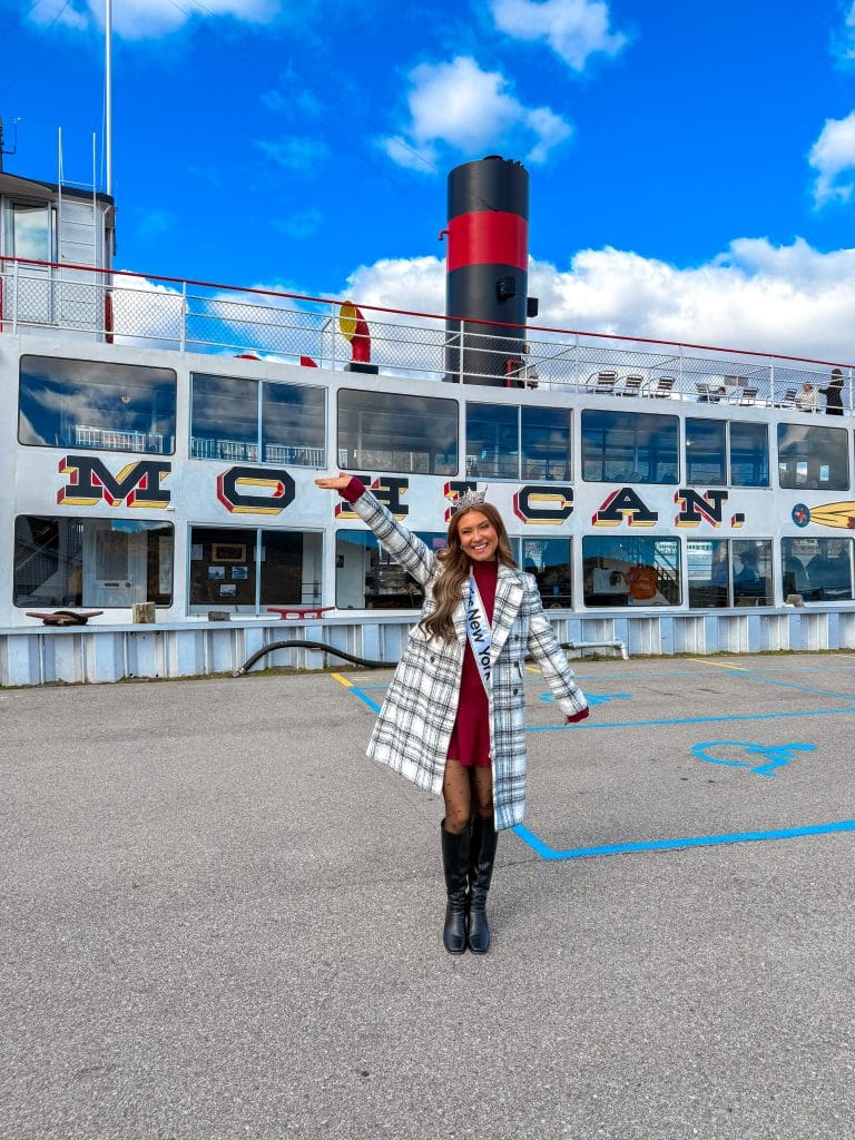 A woman in a plaid coat, maroon dress, and black boots stands smiling and waving in front of the large white Miss New York Lake George boat named Mohican under a bright blue sky with scattered clouds.