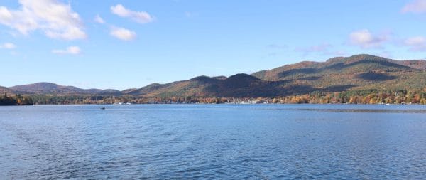 A wide, calm lake with gentle ripples, surrounded by tree-covered hills displaying autumn colors under a clear blue sky with a few scattered clouds.