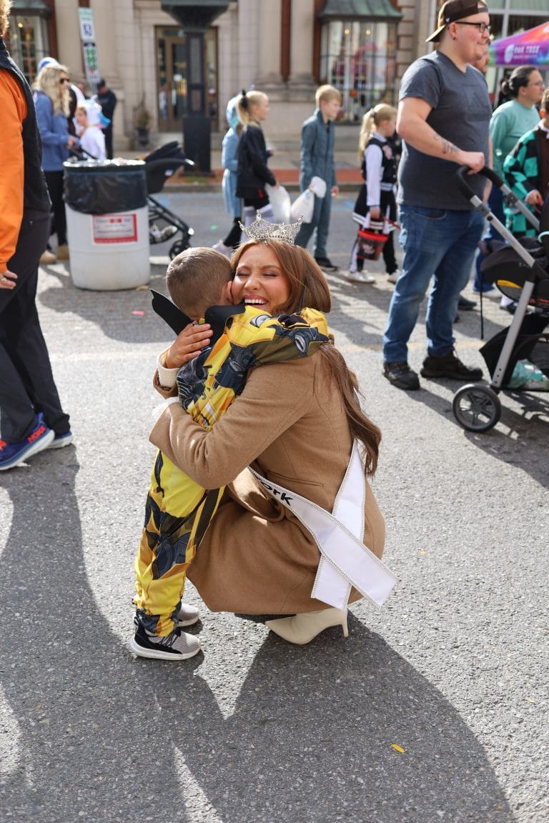 Miss New York Lake George, wearing a sash and crown, kneels and hugs a young child in a yellow costume on a busy street, surrounded by people and strollers during an outdoor event.