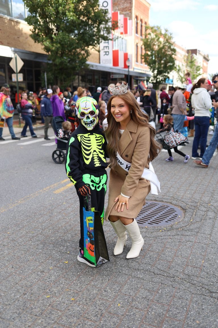 A woman in garment, possibly Miss New York Lake George, posing with a child on a lively street.