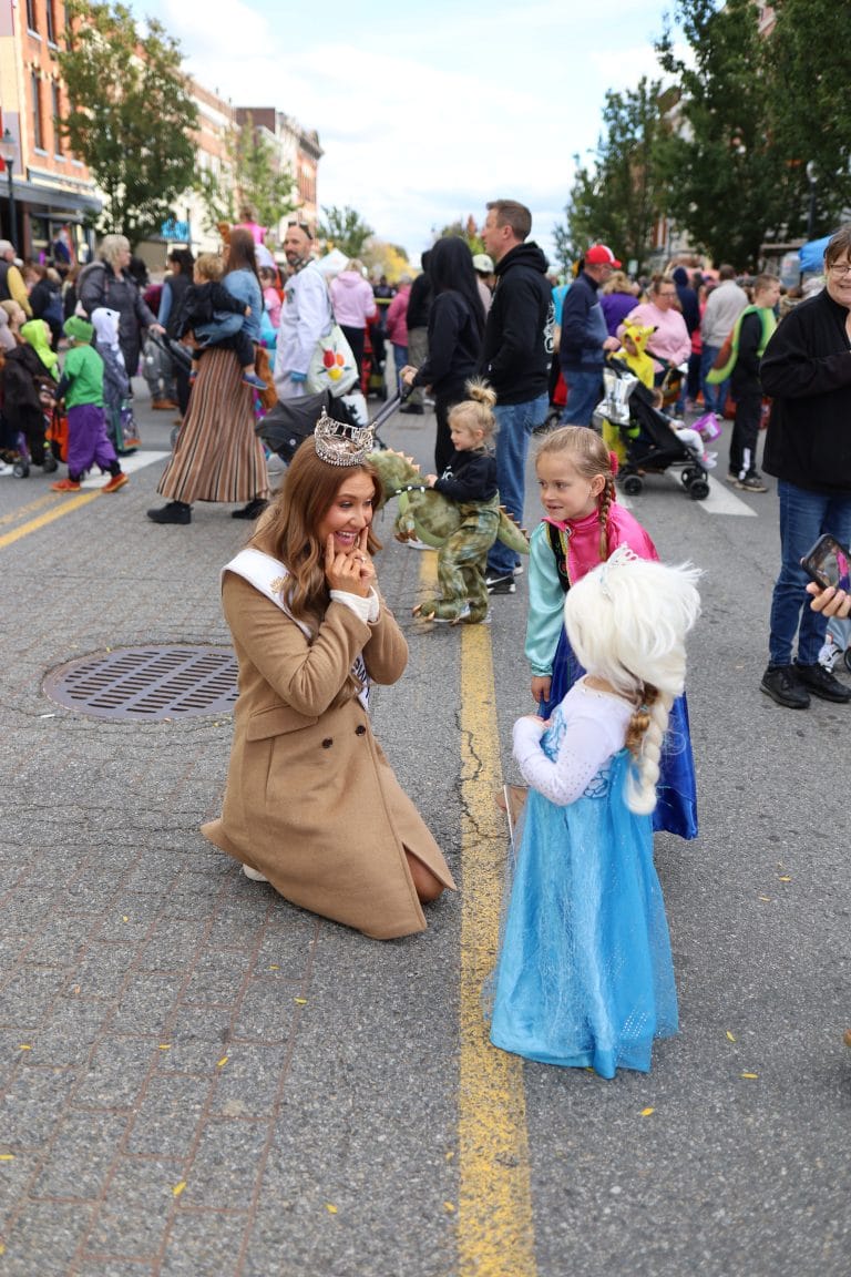 Miss New York Lake George, wearing a crown and sash, kneels on the street, smiling and chatting with two young girls dressed as princesses during a lively outdoor event filled with people in the background.