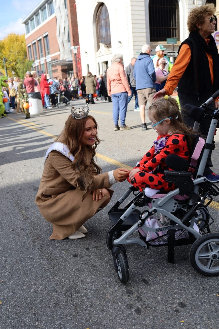Miss New York Lake George, wearing a tan coat and crown, kneels to greet a young girl in a red polka dot jacket and wheelchair at an outdoor event, with people and buildings in the background.