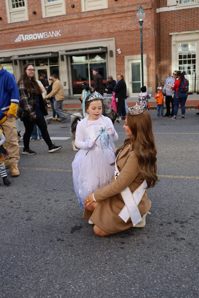 A young girl in a white dress and crown stands on a street, smiling and holding a gift while Miss New York Lake George, wearing a sash and crown, kneels in front of her during a parade. People and a bank building are visible in the background.