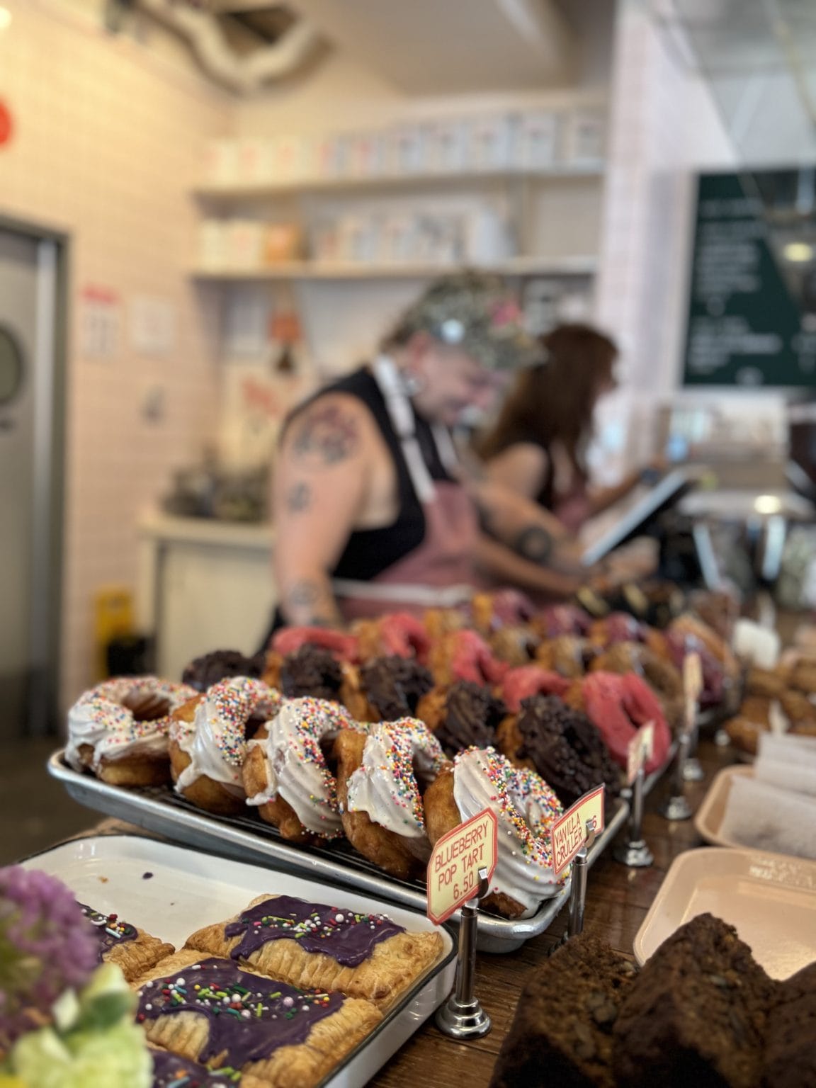 A bakery counter displays colorful donuts with sprinkles and pastries. Behind the counter, two staff members assist customers, with shelves and a menu in the background. The focus is on the vibrant baked goods.