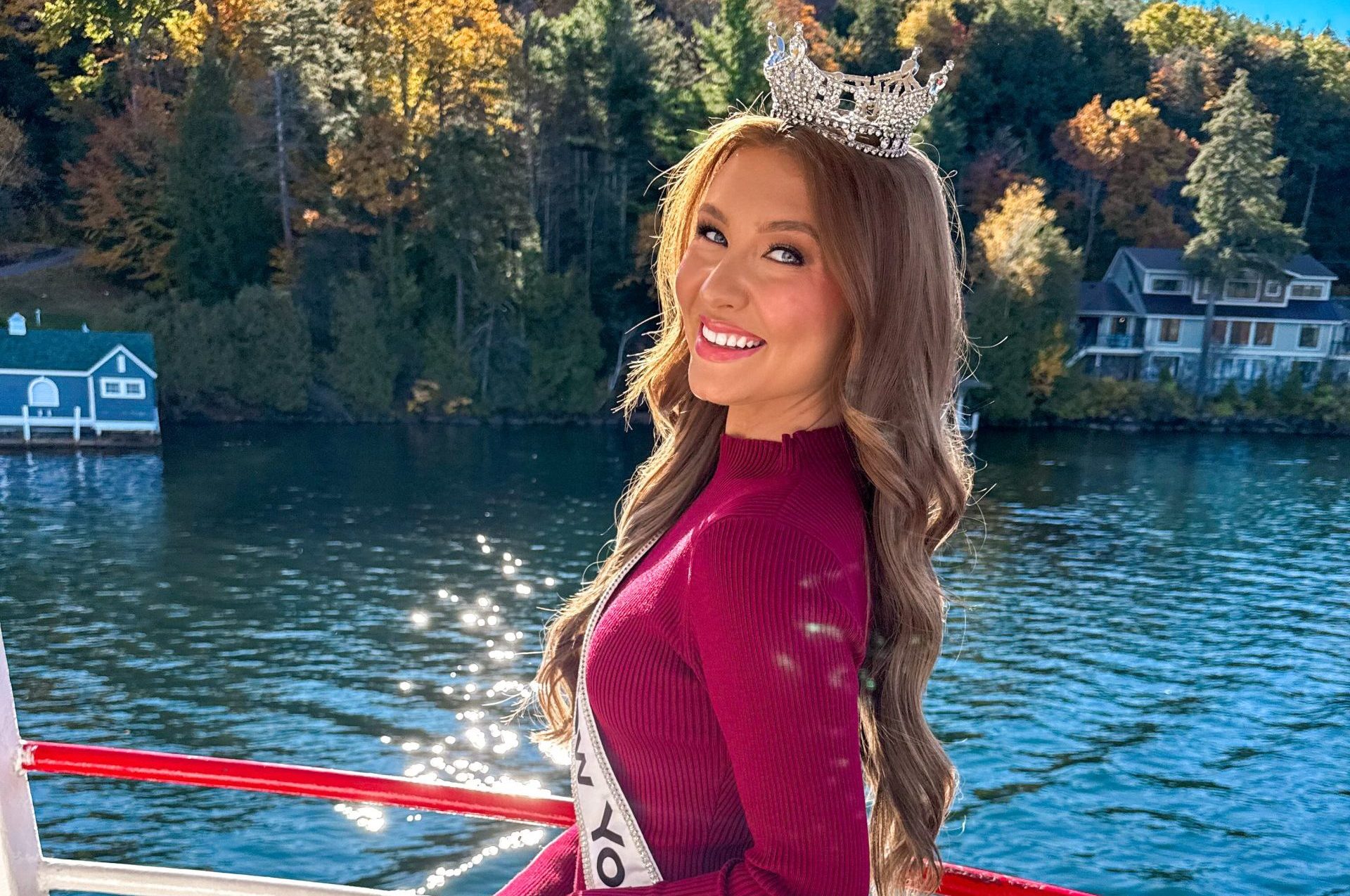 A smiling woman, Lauren Norris, crowned Miss New York in Lake George, wearing a maroon dress, sash, and black boots stands on a boat with sparkling water and vibrant autumn trees and houses in the background.