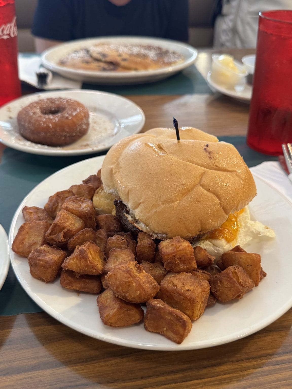 A breakfast plate with a sandwich on a bun, featuring egg and possibly sausage or bacon, served with a pile of crispy, cubed hash browns. A chocolate donut and red plastic cups are in the background on a wooden table.