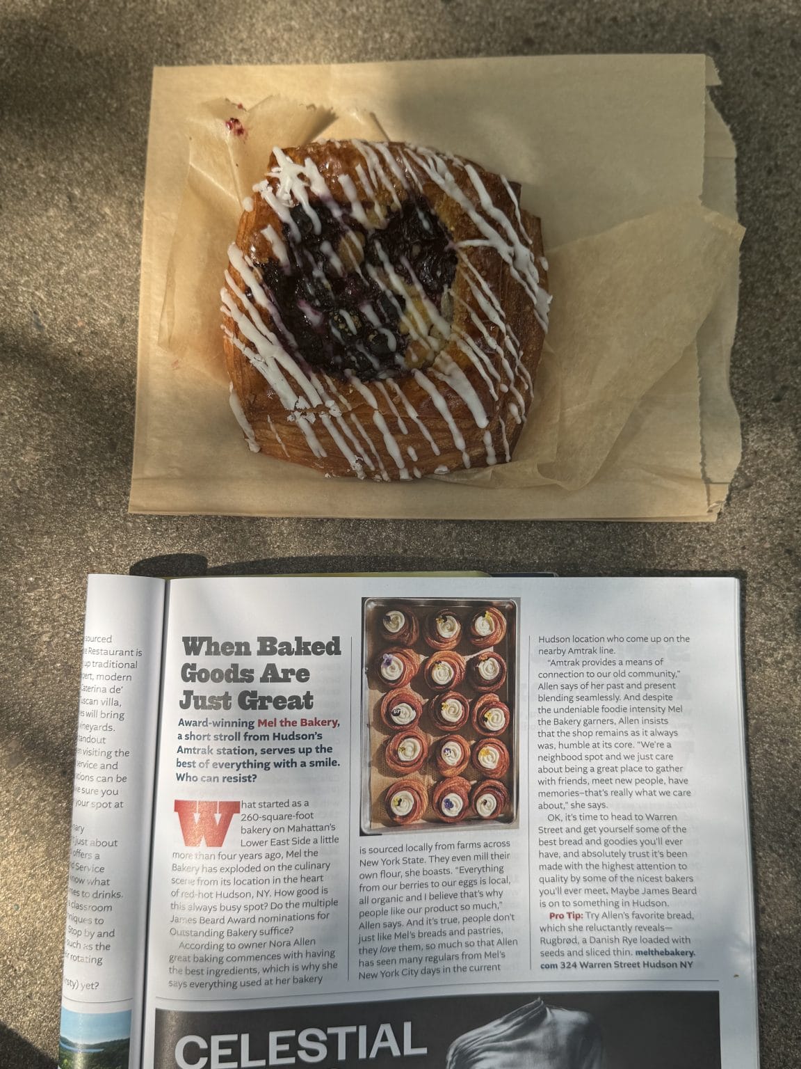 A blueberry danish with white icing sits on brown parchment paper above an open magazine featuring an article about baked goods and a photo of cupcakes. Bright natural light casts shadows on the scene.