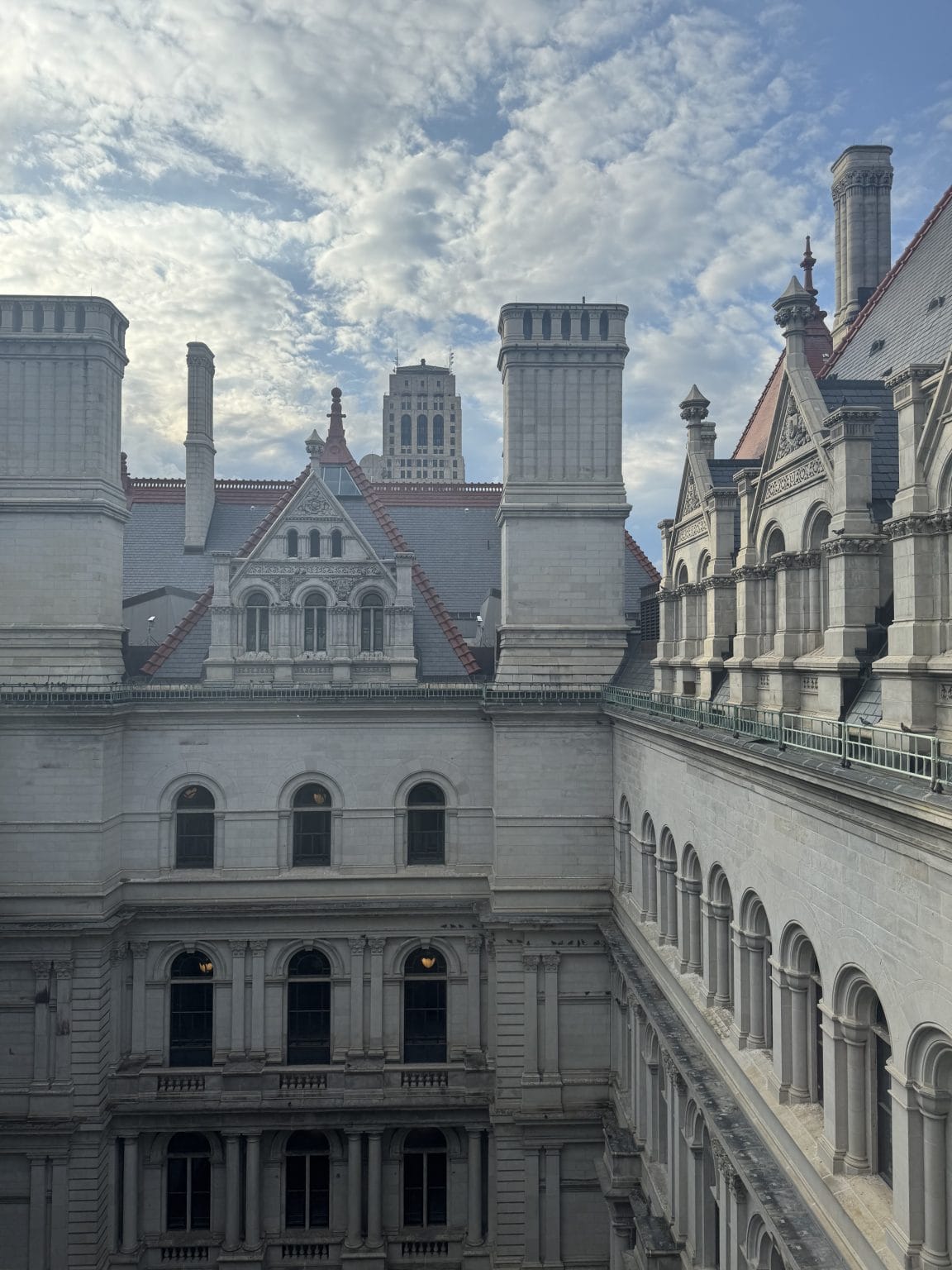 A view of a historic, ornate stone building with tall chimneys and arched windows under a partly cloudy sky; a taller skyscraper stands in the background.
