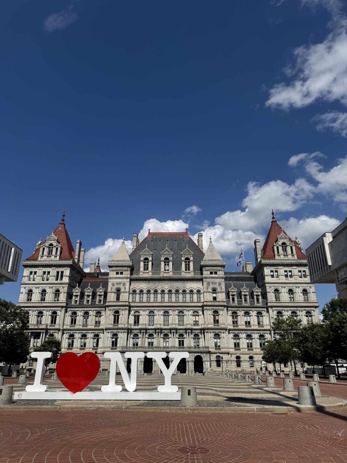 A large historic building with red-roofed towers stands under a blue sky with scattered clouds. In front, a sign reads I ♥ NY in bold white letters with a red heart.