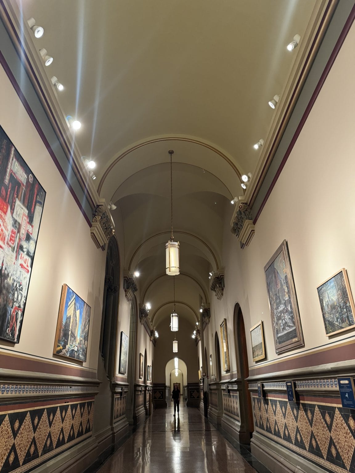 A long, elegant hallway with arched ceilings, hanging lights, and framed artwork on cream-colored walls. Two people walk in the distance, and the polished floor reflects the overhead lights.