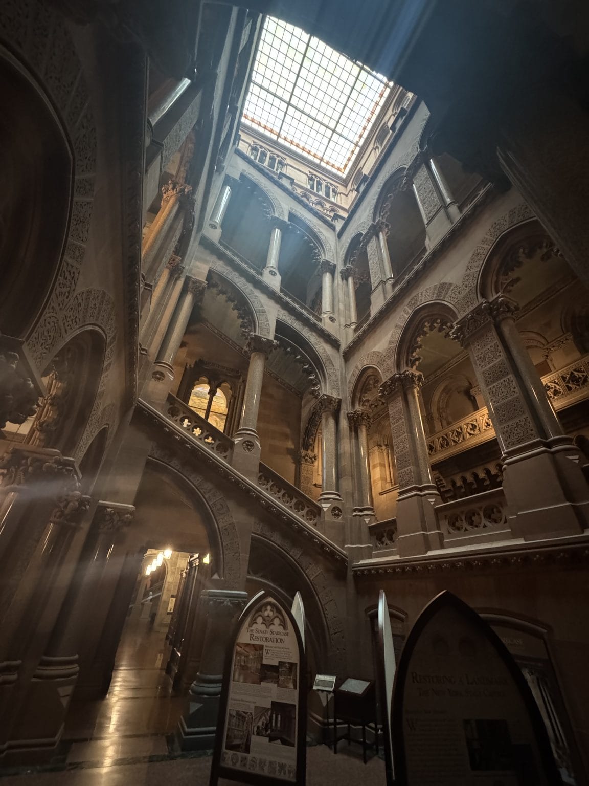 A grand, ornate interior with arched balconies, decorative columns, and a glass ceiling letting in natural light; informational displays stand on the floor below.