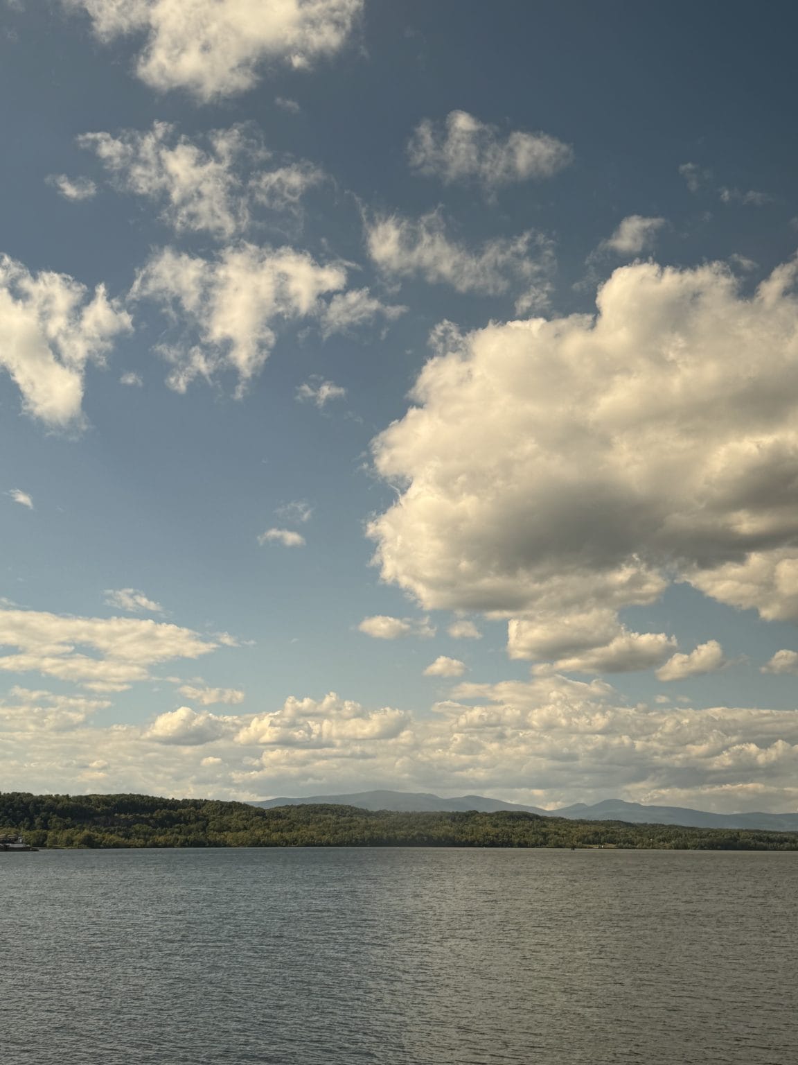 A calm lake stretches out toward a distant tree-covered shoreline under a sky partly filled with fluffy white clouds and patches of blue. Low mountains are faintly visible in the background.