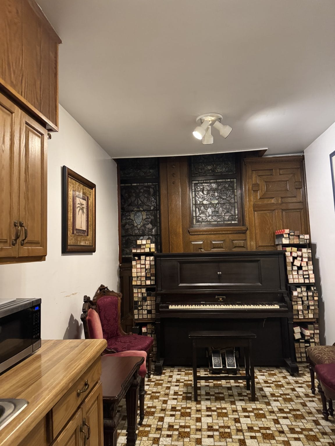 A room with a brown upright piano against a wooden and stained glass wall, flanked by stacks of piano rolls. Two ornate red chairs, a microwave, and wooden cabinetry are also visible. The floor is patterned tile.