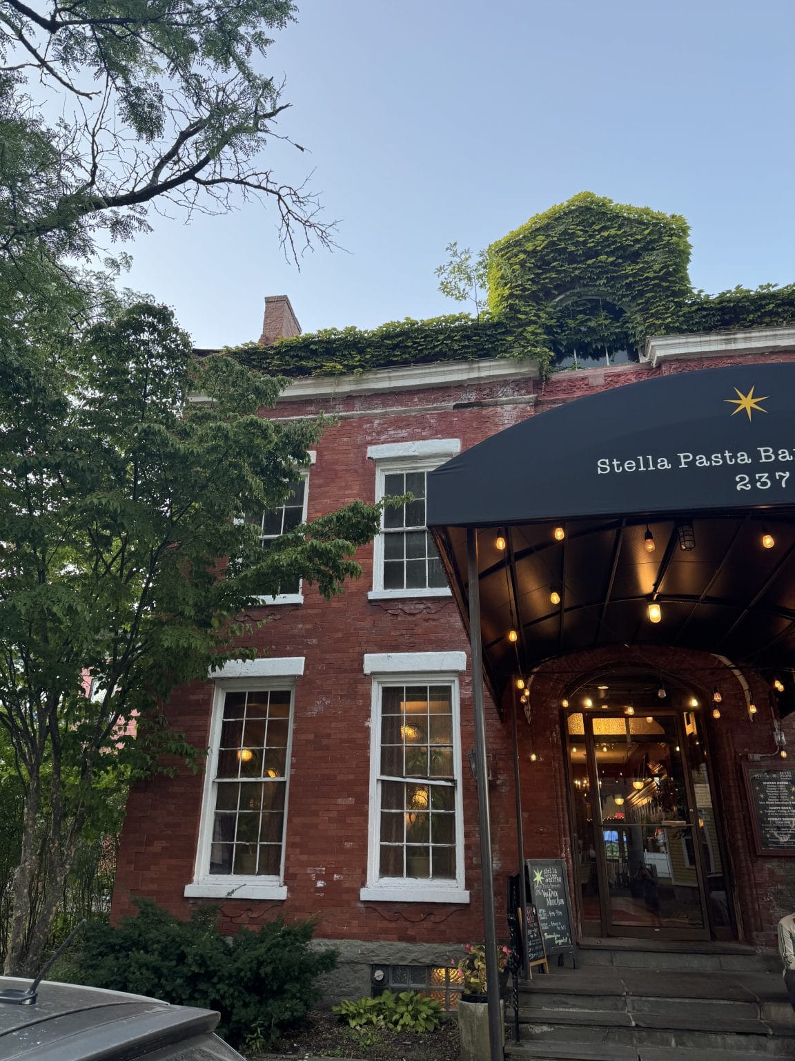 A red-brick building with large windows and ivy growing on the roof, featuring an awning reading Stella Pasta Bar 237. Warm lights glow inside and outside by the restaurant entrance. Trees frame the scene.