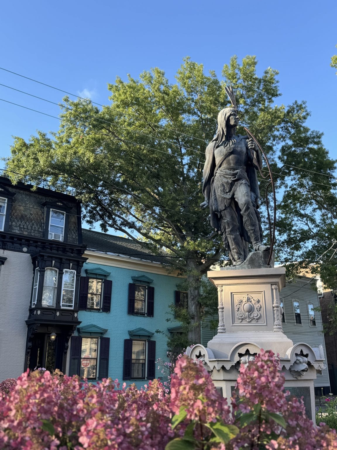 A bronze statue of a Native American figure holding a bow stands on a stone pedestal, surrounded by pink flowers, with colorful historic houses and green trees in the background under a clear blue sky.