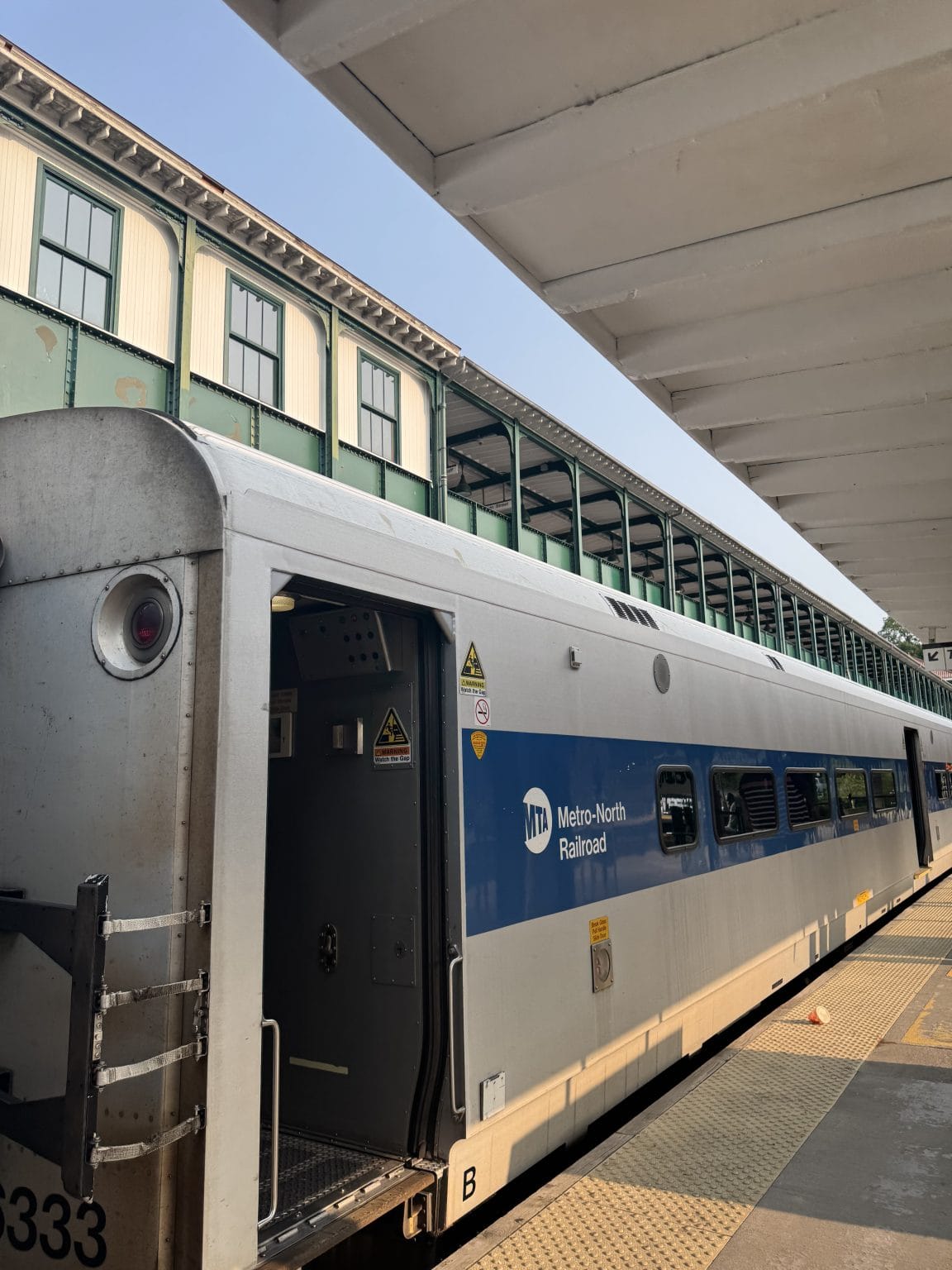 A Metro-North Railroad train waits at a sunlit outdoor station platform, with part of a green and white station building visible in the background.
