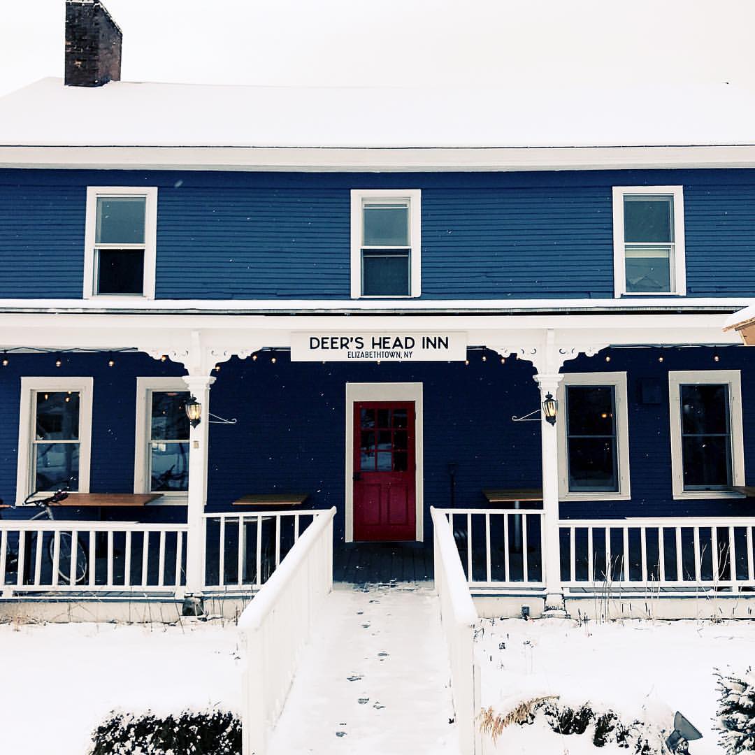 A blue two-story building with white trim, snow covering the ground and railing. The Deer's Head Inn features a red door centered under its sign, with two benches and lanterns adorning the porch.