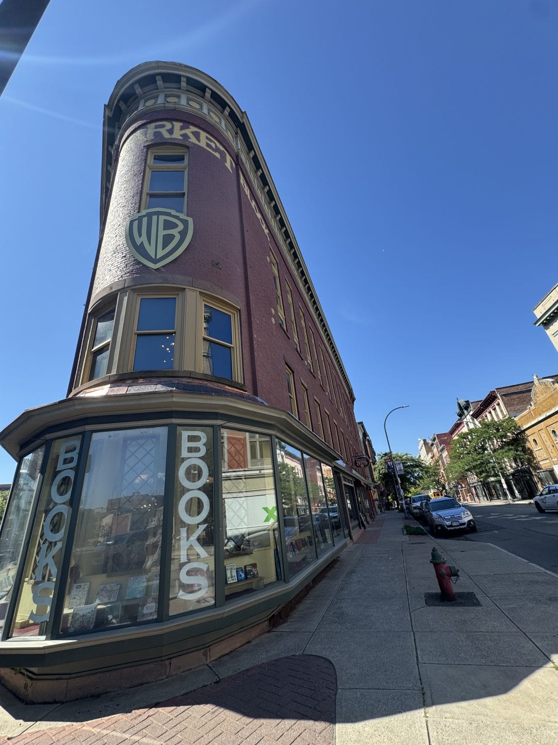 A tall, narrow brick building on a city street corner houses a bookstore with large windows displaying books. BOOKS is spelled vertically on the glass. A fire hydrant and parked cars line the sidewalk under a clear blue sky.