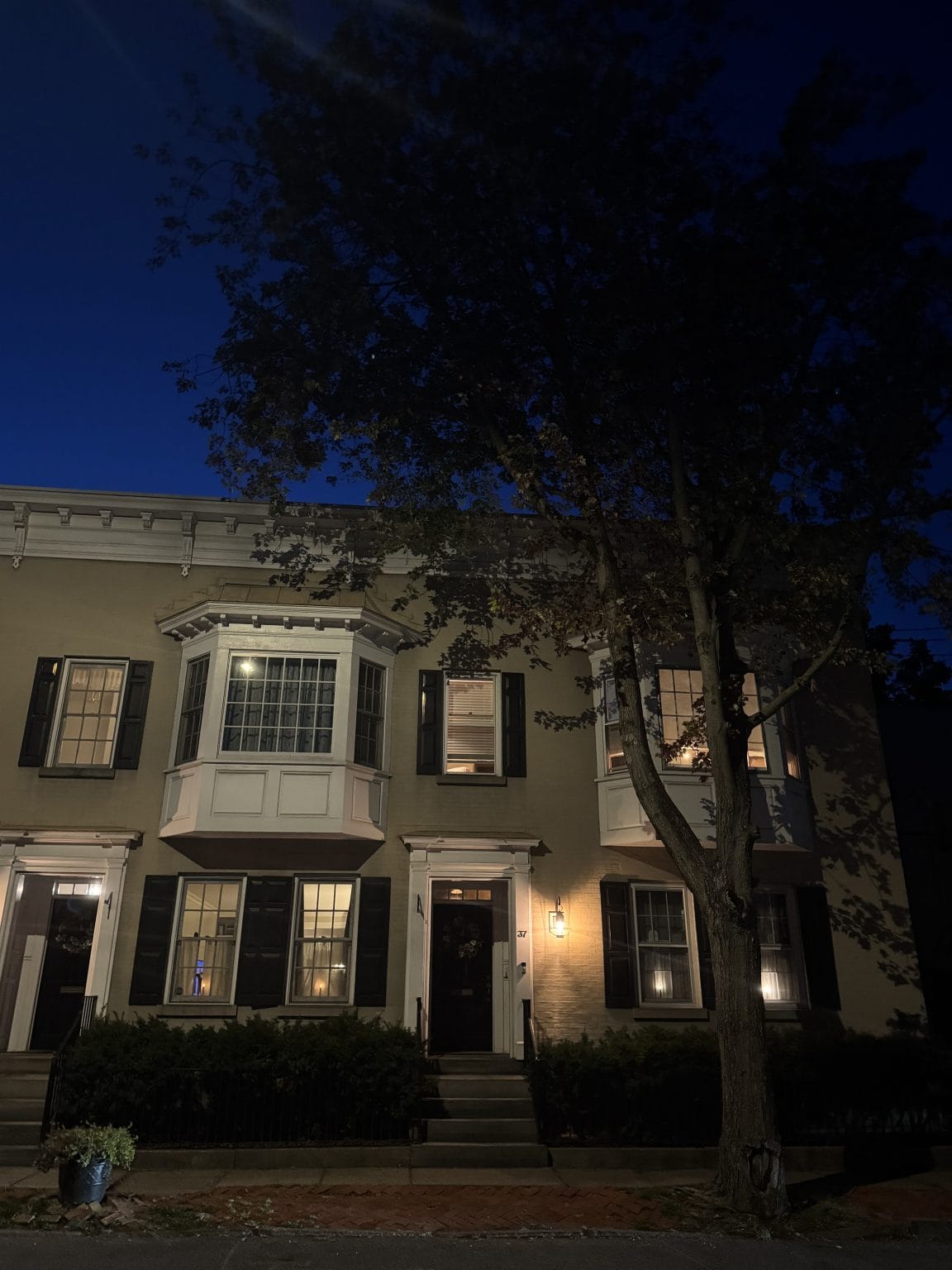 A yellow townhouse with white trim and black shutters is lit warmly from inside at night. A large tree partially obscures the house, and a blue flower pot is placed near the entrance steps. The sky is dark blue.