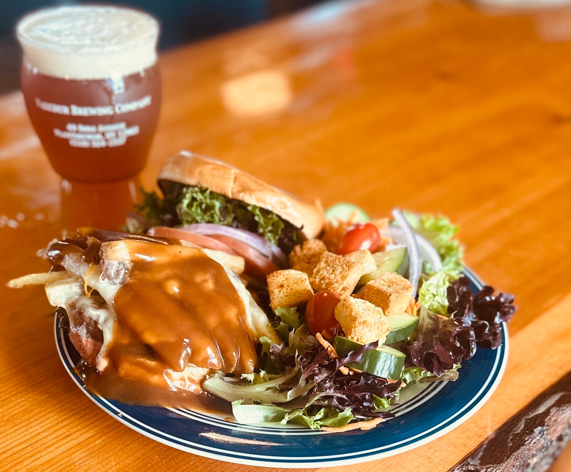 A plate from Valcour Brewing Company holds a burger, fries covered in brown gravy, and a side salad with croutons and cherry tomatoes, all sitting on a wooden table next to a refreshing glass of beer.