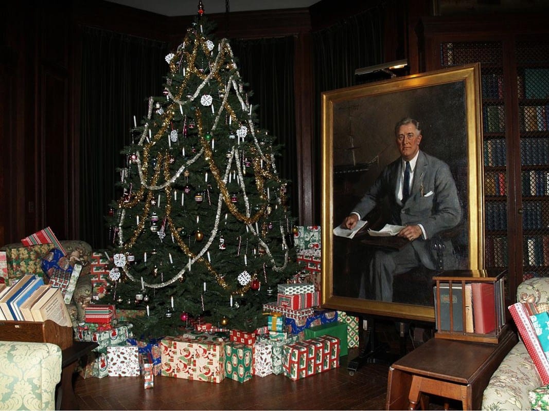 A decorated Christmas tree with tinsel and ornaments stands beside a large portrait of a man holding papers. Wrapped presents are stacked under the tree and around the portrait in a cozy, wood-paneled room.